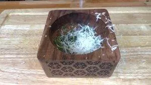 Wooden mortar with shredded cheese and herbs on a cutting board. Perfect for pesto or seasoning preparation.