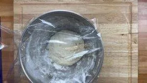 Bread dough rising in a covered bowl on a wooden surface, ready for baking.