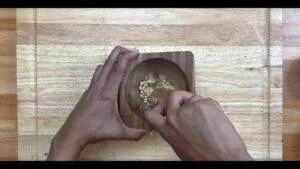 Person crushing pine nuts in a wooden mortar with a pestle on a wooden surface.