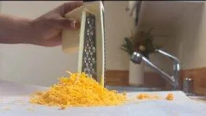 Hand grating cheddar cheese in a kitchen, with a sink and plant visible in the background.