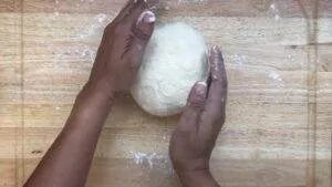 Hands kneading round dough on a floured wooden board, preparing for baking.