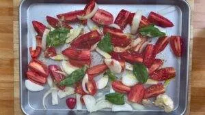 Sliced tomatoes, onions, garlic, and basil with olive oil on a baking sheet, ready for roasting.
