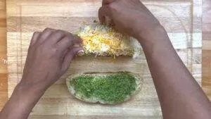 Preparing cheese and pesto sandwich on wooden board. Hands adding shredded cheese on bread with green pesto spread.