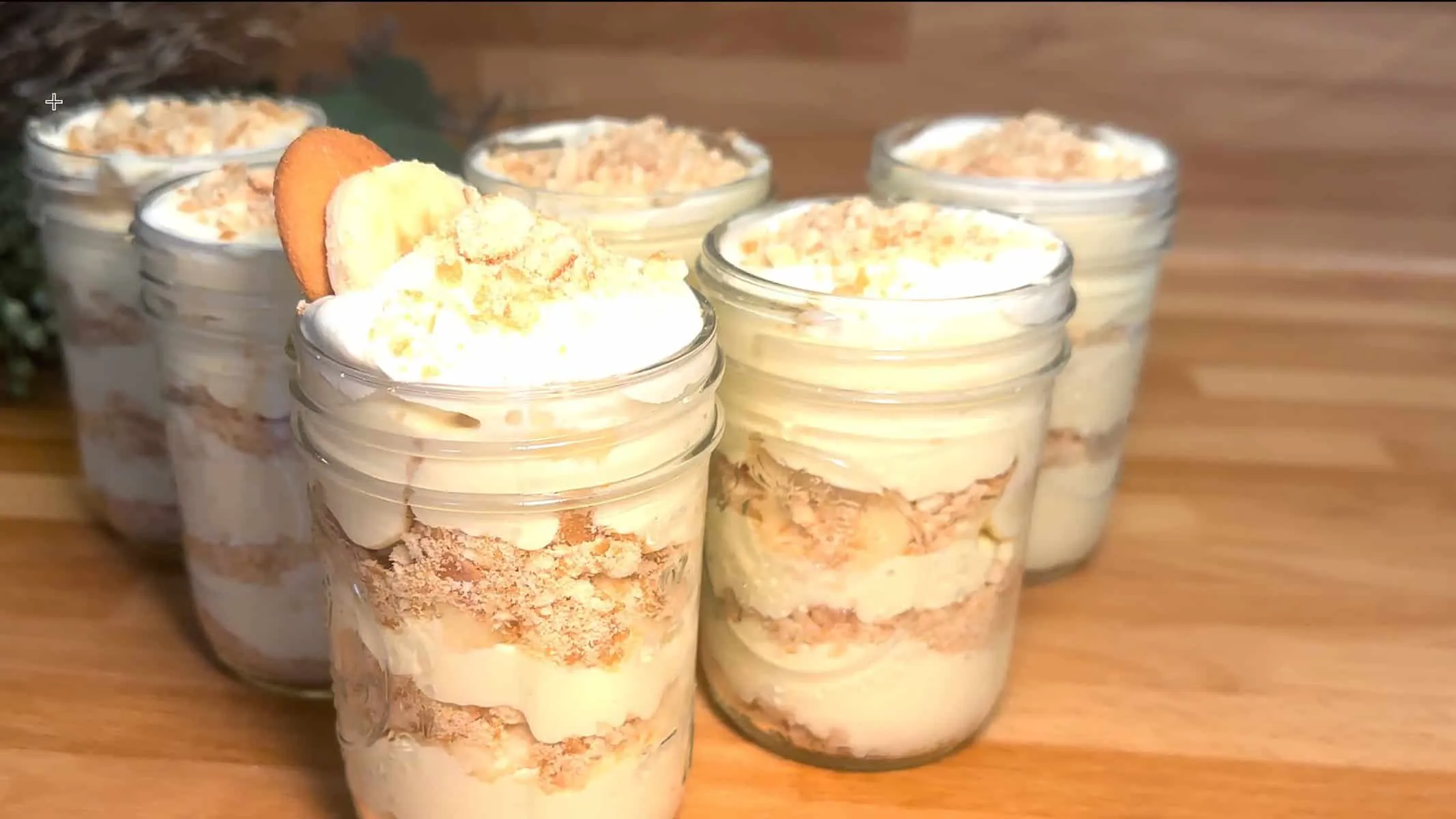 Banana pudding parfaits with whipped cream and crushed cookies in mason jars on a wooden table.