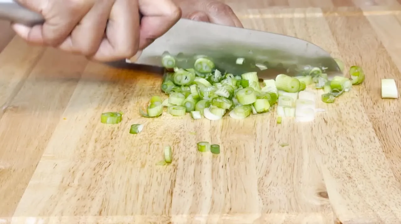 Hand chopping green onions on a wooden cutting board with a sharp knife.
