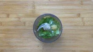 Basil, garlic, and oil in a food processor on a wooden board, ready to make fresh pesto sauce.