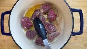 Cooking beef chunks in a pot with tongs for a savory stew on a wooden surface.