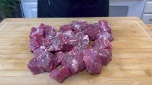Raw beef cubes seasoned with salt on a wooden cutting board, ready for cooking.