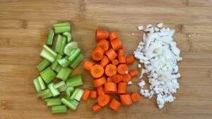 Chopped celery, carrots, and onions on a wooden cutting board, ready for cooking.