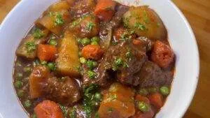 Hearty beef stew with potatoes, carrots, and peas in rich gravy served in a white bowl, topped with fresh herbs.