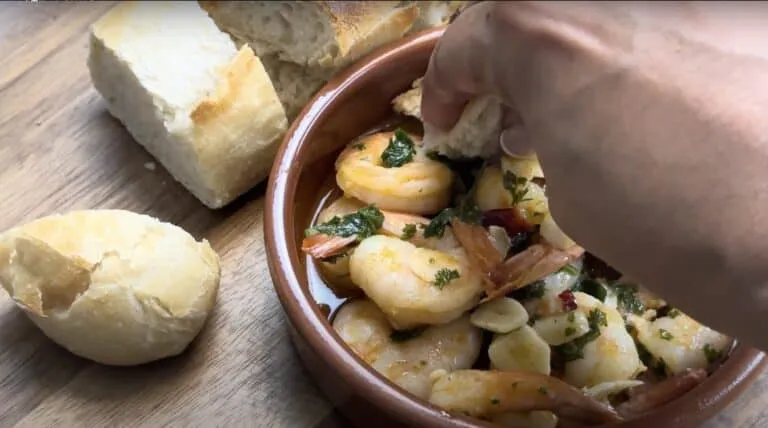 Hand dipping bread into a bowl of Gambas Pil Pil with fresh herbs; served with crusty bread on a wooden table.