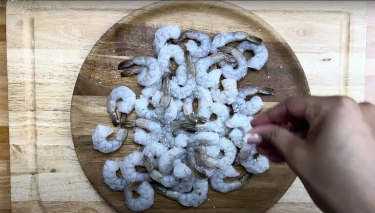 Raw shrimp being seasoned with salt on a wooden plate.