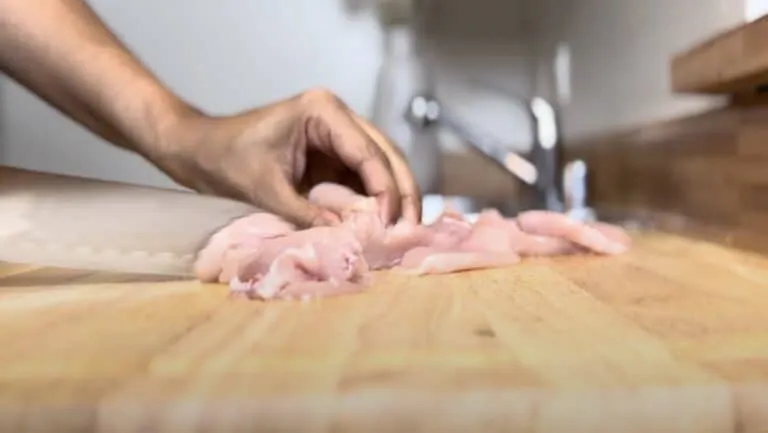 Person slicing raw chicken on a wooden cutting board in a kitchen setting.