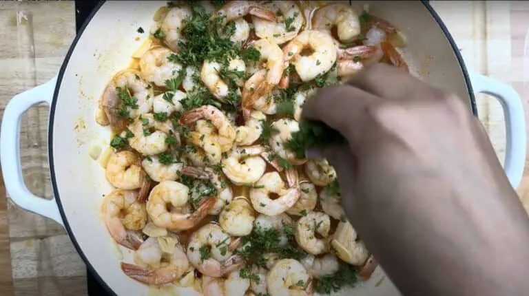 Person garnishing Gambas Pil Pil with herbs in a white pan. Perfect for seafood recipe inspiration.