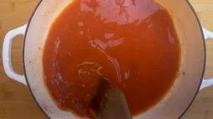 Simmering tomato sauce in a white pot, stirred with a wooden spoon on a wooden countertop.