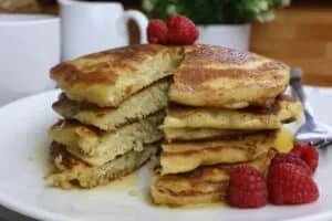 Stack of fluffy pancakes with syrup and raspberries on a white plate, perfect breakfast treat.