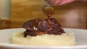 Slow-cooked short ribs with gravy over mashed potatoes on a white plate.