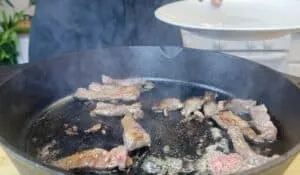 Sizzling meat strips cooking in a cast iron pan, ready to be served on a white plate, with smoke rising.