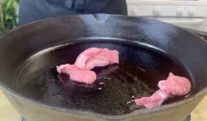 Raw beef slices sizzling in a hot cast iron pan on a stovetop.