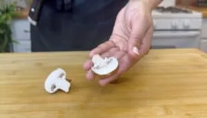 Person holding a sliced mushroom over a wooden cutting board in a kitchen setting.