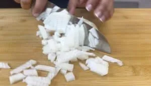 Chopping white onion on a wooden cutting board with a sharp knife in a kitchen setting.