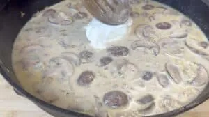 Creamy mushroom sauce in a skillet with wooden spoon stirring sour cream.