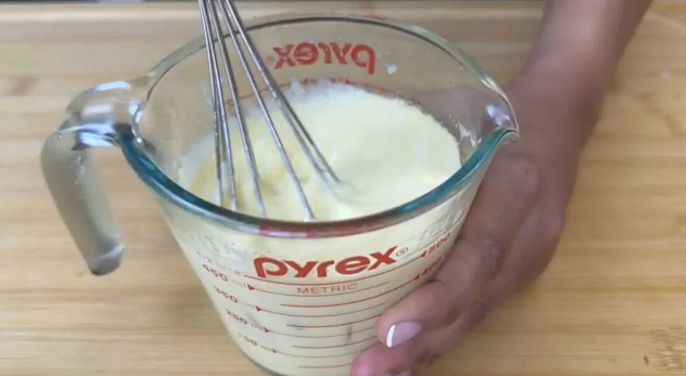 Whisk mixing creamy batter in Pyrex measuring cup on wooden surface.