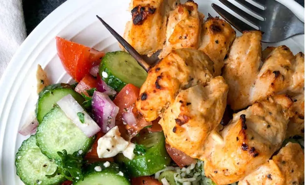 Grilled chicken skewers with a fresh cucumber tomato salad on a white plate.