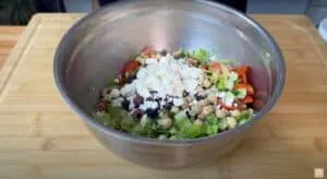 Fresh Greek salad in a metal bowl with feta, cherry tomatoes, lettuce, cucumbers, and chickpeas on a wooden board.