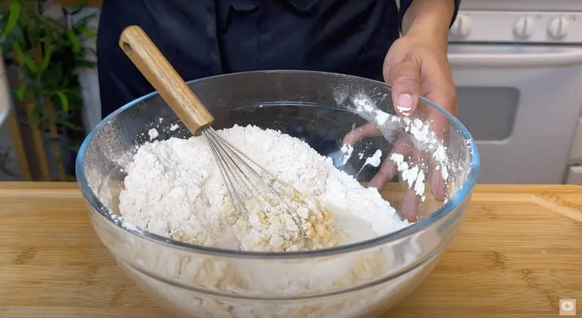 Person mixing flour and liquid in a clear bowl with a whisk on a wooden table.