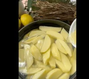 Sliced potatoes soaking in water with fresh lemons and herbs in the background. Perfect for a refreshing recipe.