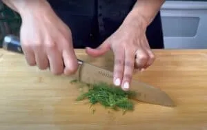 Person chopping fresh dill on a wooden cutting board with a knife.