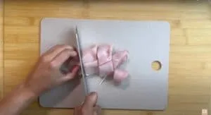 Hands chopping raw chicken on a cutting board for cooking preparation.