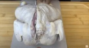Raw chicken being cut in half for spatchcocking on a cutting board.