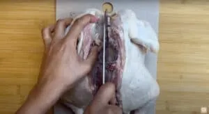 Person spatchcocking a chicken on a cutting board with a knife; preparing poultry for cooking.