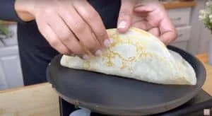 Person folding a freshly cooked crepe on a non-stick pan in a kitchen setting.