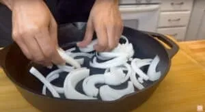 Hands arranging sliced onions in a cast iron skillet on a wooden countertop.