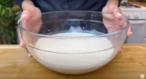 Person holding a clear glass bowl of pancake batter on a wooden countertop.