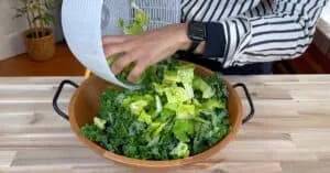 Person adding fresh greens from salad spinner to a wooden bowl on a kitchen countertop.