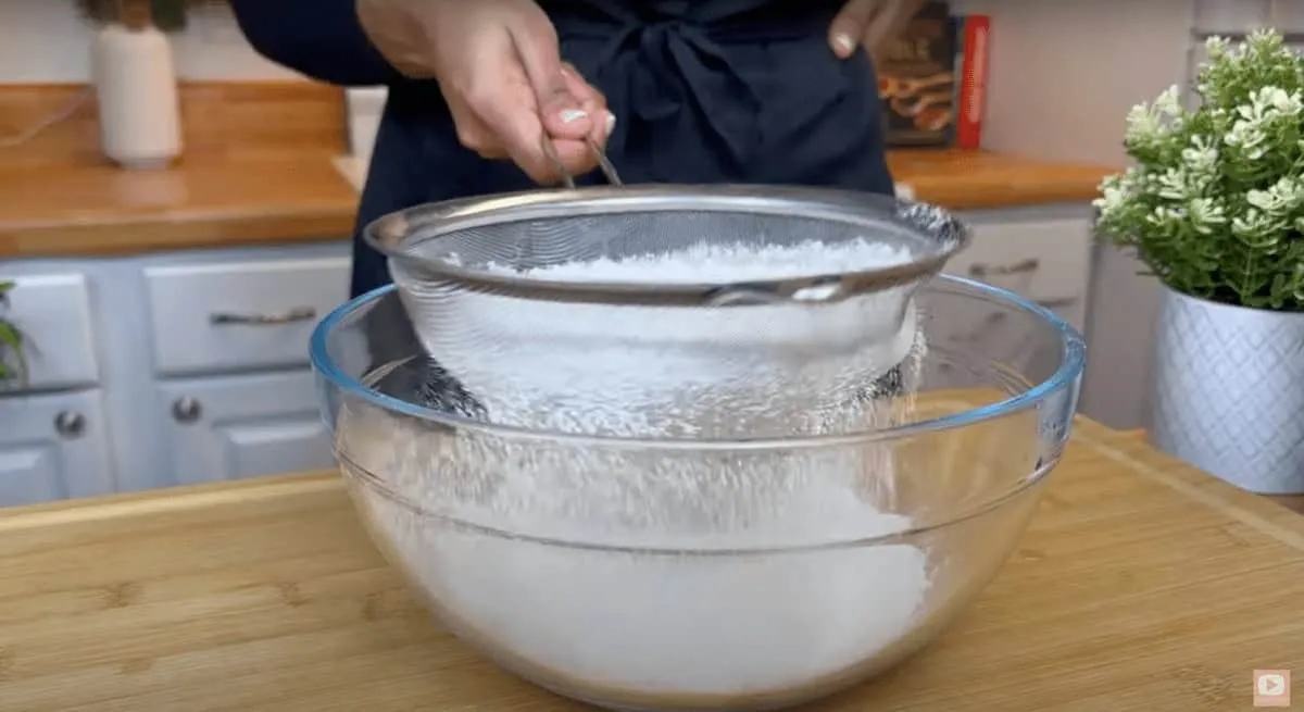 Person sifting flour into a glass bowl on a wooden countertop in a home kitchen.