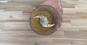 Ground ingredients in a food processor bowl on a wooden countertop, hand holding the container.