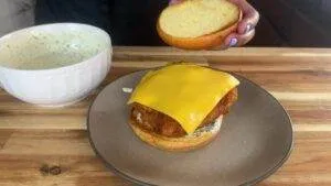 Chicken sandwich with cheese and sauce being prepared, next to a bowl of creamy sauce on a wooden table.