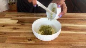 Person adding chopped pickles to a bowl with mashed potatoes on a wooden kitchen counter.
