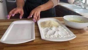 Hands preparing breading station with flour, breadcrumbs, and egg wash in a kitchen setting.