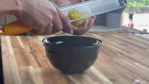 Person zesting a lemon over a black bowl on a wooden countertop for cooking preparation.