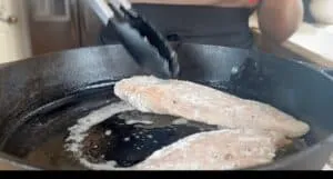 Cooking breaded chicken fillets in a cast iron skillet with tongs.