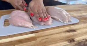 Person slicing raw chicken breasts on a cutting board with bright red nail polish.