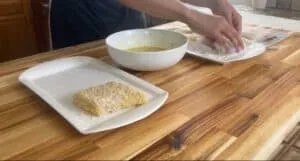Person breading fish fillet with flour and egg on wooden kitchen counter.