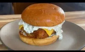 Crispy chicken sandwich with cheese and creamy dill sauce on a fresh bun served on a plate.