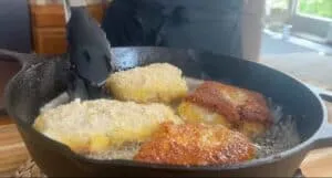 Fried breaded fish fillets sizzling in a cast iron skillet with tongs. Perfect for crispy seafood recipes.